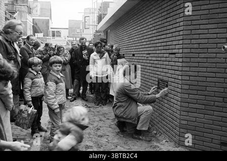 Nuove case di Tuinstraat Eerste Steen Streets Haarlem Tuinstraat Paesi Bassi, Whizgle News, Dutch Desk, Paesi Bassi, 1950 - 2000 su 11-11-1983. L'immagine mostra questi argomenti. La scena cattura un raduno di persone in un ambiente all'aperto, sullo sfondo di un muro di mattoni che viene costruito o svelato. La parete è composta da un ricco mattone marrone-rossastro, che mostra una struttura ordinata e ordinata che attira l'attenzione. In primo piano, una persona si inginocchia vicino al muro, concentrandosi intensamente su un oggetto che sta posizionando o fissando alla superficie. Questo individuo è vestito di casua Foto Stock