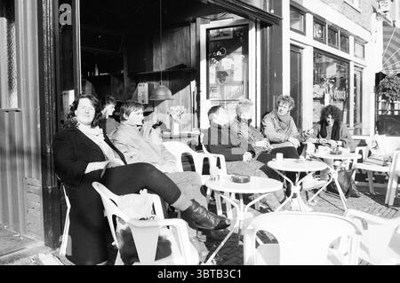 Persone che prendono il sole mercato del burro prendere il sole Haarlem Botermarkt Paesi Bassi, Whizgle News, Dutch Desk, Paesi Bassi, 1950 - 2000 il 10-03-1987. L'immagine mostra questi argomenti. In una vivace scena di strada, un gruppo di sette persone occupa un'accogliente area all'aperto in un bar. Si siedono intorno a piccoli tavoli rotondi, ciascuno decorato con una tazza, suggerendo conversazioni informali o momenti di riflessione. Le persone sono un mix di uomini e donne, di aspetto e di comportamento diversi. Il loro abbigliamento riflette un'atmosfera rilassata e urbana, con alcune giacche indossate e altre in un abbigliamento casual. La donna all'estrema sinistra, arguzia Foto Stock