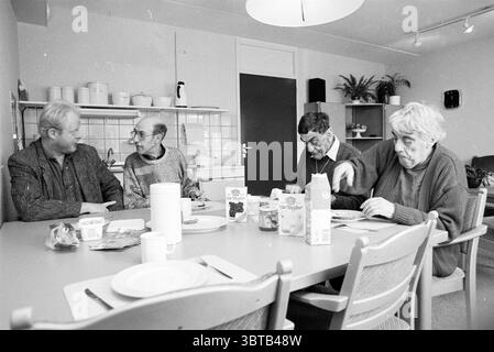 Boerlage House IJmuiden Interior IJmuiden Paesi Bassi, Whizgle News, Dutch Desk, Paesi Bassi, 1950 - 2000 il 13-03-1990. L'immagine include questi argomenti. In una cucina modesta e ben illuminata, quattro persone si siedono intorno a un tavolo circolare fatto di legno chiaro. La camera ha un'atmosfera calda e invitante, illuminata da un'illuminazione soffusa. Ogni persona contribuisce a una scena di interazione silenziosa. Da un lato, due uomini con gli occhiali si impegnano in conversazione. Uno ha un'acconciatura di colore chiaro e leggermente ruvida e sfoggia un maglione scuro e testurizzato. L'altro, con una costruzione più sottile, ha baffi e d Foto Stock