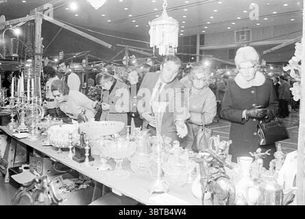 Mercato dell'antiquariato Hillegom Markt, Whizgle News, Dutch Desk, Paesi Bassi, 1950 - 2000 il 07-11-1971. Questi argomenti sono mostrati nell'immagine. La scena cattura un vivace mercato al coperto pieno di una vasta gamma di oggetti d'epoca e di antiquariato esposti su lunghi tavoli. I tavoli sono rivestiti di vetreria, argenteria e vari oggetti decorativi, che riflettono una ricca varietà di stili ed epoche. Vasi di vetro lucenti, candelabri decorati e figure intricate attirano l'attenzione, mettendo in risalto la loro maestria artigianale. Un gruppo di persone sta in piedi insieme, le loro espressioni un mix di curiosità ed eccitazione. Quindi Foto Stock