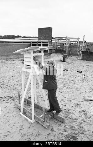 Capanna termometrica servizi meteo, Whizgle News, Dutch Desk, Paesi Bassi, 1950 - 2000 il 07-12-1964. L'immagine include questi argomenti. In un ambiente sabbioso all'aperto, un uomo è impegnato nella costruzione di un'alta struttura bianca che assomiglia a una torre di osservazione o di osservazione. Si trova su una piattaforma in legno improvvisata, posizionando con cura ulteriori spranghe in legno sulla parte superiore del telaio. L'individuo è vestito con un blazer scuro, con pantaloni di colore chiaro, che trasuda un senso di determinazione e maestria mentre si concentra sul suo compito. La sabbia intorno a lui è beige chiaro, con una consistenza leggermente grossolana Foto Stock