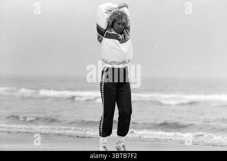 Donna che fa esercizio sulla spiaggia., Whizgle News, Dutch Desk, Paesi Bassi, 1950 - 2000. Questi argomenti sono mostrati nell'immagine. In una scena costiera, l'attenzione è rivolta a un individuo in piedi sulla spiaggia sabbiosa. Sono caratterizzati da un outfit sportivo, con una giacca dai colori chiari a righe audaci e pantaloni sportivi aderenti che si rastremano alle caviglie. Il tessuto appare leggero, perfetto per l'atmosfera ventilata. I capelli della persona sono modellati con onde morbide, dando un'impressione spensierata, e sono impegnati in un esercizio di stretching, suggerendo uno stile di vita attivo. Lo sfondo mostra un'ampia Foto Stock