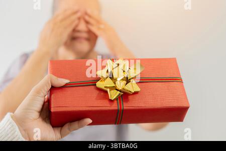 Primo piano di una scatola regalo rossa di natale tenuta per mano della figlia per una sorpresa da regalare alla sua anziana madre. Concetto di offerta a sorpresa. Foto Stock
