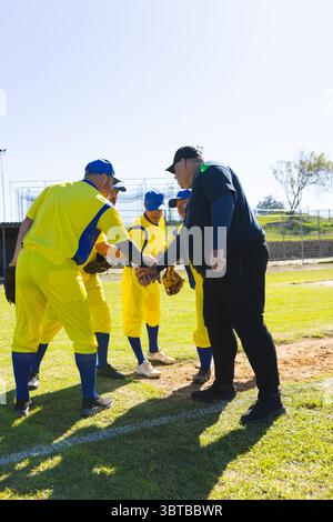 Giocatori di baseball e allenatore maschili che si accoccolano sul campo al tumulo, mettendo le mani in uniforme e guanti Foto Stock