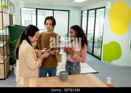 Diversi colleghi in piedi intorno a un tavolo di legno che reggono tazze da caffè che rivedono lo smartphone nella sala d'attesa Foto Stock