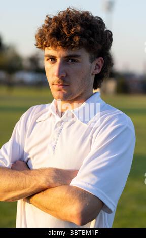 Uomo in piedi con le braccia incrociate sul campo sportivo durante il tramonto indossando una polo con orecchini a cerchio Foto Stock
