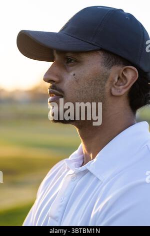 Uomo indiano in piedi sul campo al tramonto con berretto da baseball nero e polo, copia spazio Foto Stock