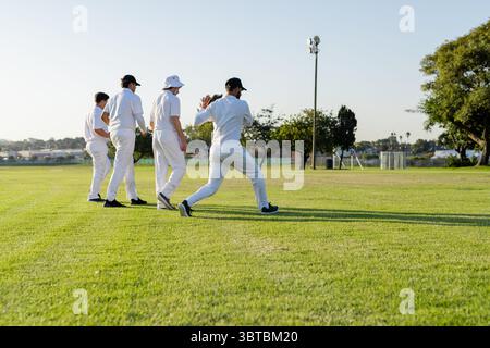 Diversi compagni di squadra maschili che camminano in un'unica fila attraverso il campo di cricket indossando divise bianche alla luce del sole Foto Stock