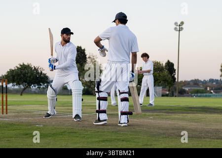 Diversi compagni di squadra che giocano a cricket su un terreno sportivo di periferia indossando una divisa bianca e tenendo in mano pipipistrelli Foto Stock