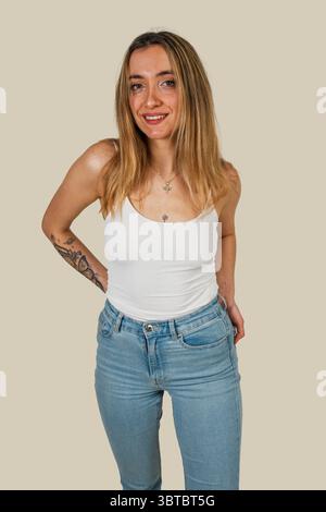 Ritratto in studio di una giovane donna allegra con tatuaggi che indossano una canotta bianca e jeans, che mostra uno stile alla moda e sicuro di sé Foto Stock