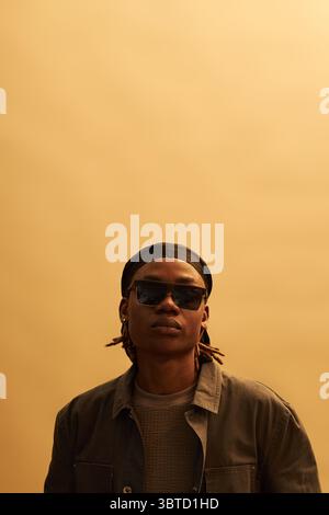 Ritratto di un giovane uomo nero adulto che indossa occhiali da sole su uno sfondo semplice, che guarda direttamente la fotocamera con un'espressione neutra, acconciati in dreadlock corti Foto Stock