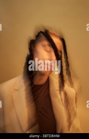 Ritratto di donna nera giovane adulta con trecce lunghe che inclinano la testa e chiudono gli occhi, effetto di sfocatura del movimento che crea uno sfondo dinamico e neutro che enfatizza il soggetto Foto Stock