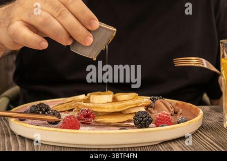 Versare a mano lo sciroppo su una pila di pancake conditi con fette di banana e frutti di bosco misti, creando un'atmosfera accogliente per la colazione piena di calore e sapore Foto Stock