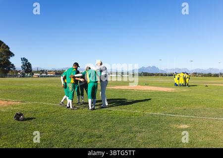 Giocatori di baseball e allenatore maschili si accoccolano sul tumulo del lanciatore sul campo da baseball con il casco del ricevitore Foto Stock