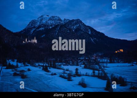 Vista aerea del castello di Neuschwanstein e del castello di Hohenschwangau si illuminano lentamente sullo sfondo scuro della montagna sotto un cielo crepuscolo, Schwangau, Bayern, Germania. Foto Stock