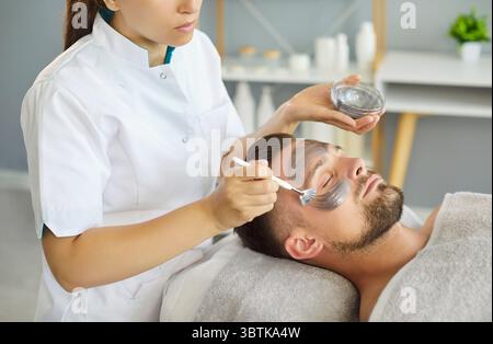 Donna estetista che applica la maschera cosmetica in argilla con un pennello sul bel viso dell'uomo nel salone termale. Foto Stock