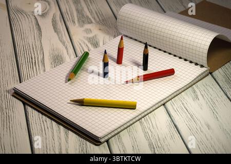 Blocco note vuoto aperto con carta a scacchi con matite colorate su un tavolo in legno bianco con sfondo didattico di cancelleria a luce crepuscolare Foto Stock