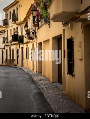 Tranquilla strada stretta costeggiata da case in stile mediterraneo tradizionale e lanterne d'epoca, catturate durante l'ora d'oro in un villaggio spagnolo. Foto Stock