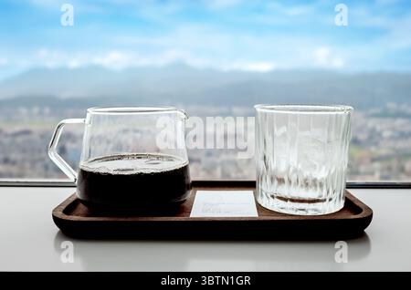 Primo piano di una caraffa di vetro trasparente di caffè nero e un bicchiere di cubetti di ghiaccio su un vassoio vicino a una finestra che si affaccia sulla città Foto Stock