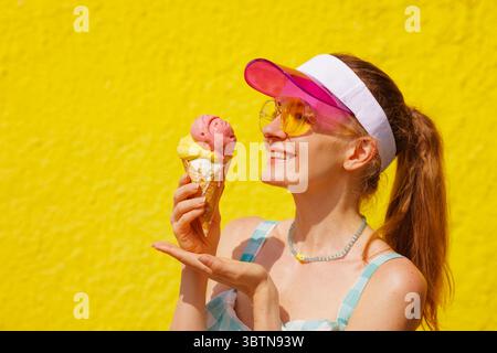 Donna sorridente in occhiali da sole che tiene in mano un cono gelato fuso all'aperto in una giornata estiva di sole, con sfondo giallo brillante Foto Stock