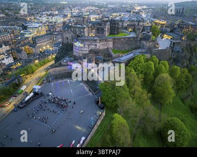 Edimburgo, Regno Unito - 16 giugno 2025: Veduta aerea del maestoso Castello di Edimburgo arroccato sulla Castle Rock, le sue antiche mura di pietra che si illuminano dolcemente nella luce che svanisce, giustapposta ai verdi vibranti degli alberi circostanti e all'arazzo urbano della città sottostante, dove piccole figure si riuniscono nella spianata, illuminata da una costellazione di luci al tramonto. Foto Stock