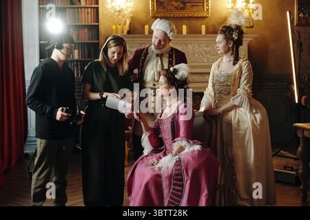 Gruppo multietnico, tra cui giovane donna adulta in abito barocco seduta e tenuta in mano la sceneggiatura, mentre giovane uomo e donna adulta in costumi rinascimentali in piedi, due membri della troupe assistono sul set del film Foto Stock