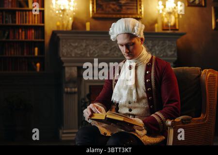 Giovane uomo adulto caucasico che indossa un costume barocco storico seduto in una stanza ornata che legge un libro, parrucca in polvere e abiti elaborati che riflettono lo stile rinascimentale Foto Stock