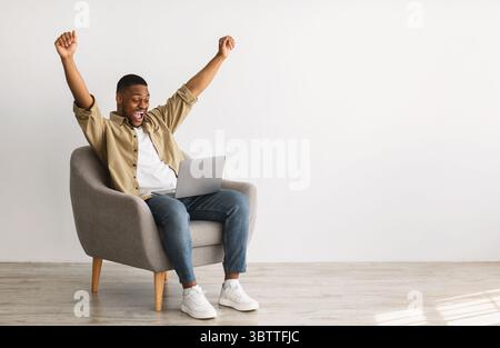 Joyful African Male Using laptop shaking Fists over Grey Wall Foto Stock