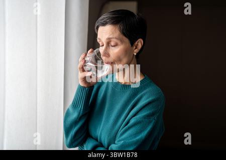 Una donna triste e tranquilla sorseggia acqua alla finestra persa in un ambiente isolato al chiuso. Tensione nervosa, calmante Foto Stock