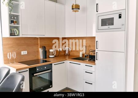 Un'elegante cucina moderna con armadi bianchi, dettagli in legno ed elettrodomestici all'avanguardia che presentano un design minimalista e organizzato con luce brillante Foto Stock