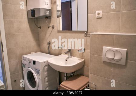 Un bagno moderno e compatto con lavatrice, lavello, scaldabagno e specchio, il tutto posto su pareti piastrellate beige, che mostrano un uso efficiente di Foto Stock