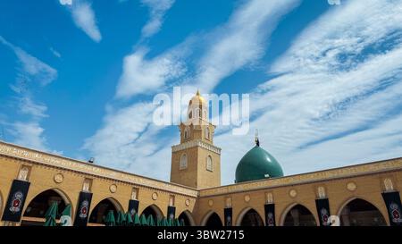 La cupola dorata e la torre si innalzano sul cortile di Kufa in Iraq, mostrando splendidi dettagli architettonici contro un cielo blu brillante. Foto Stock