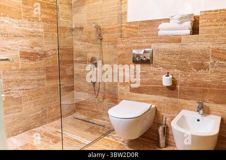 Un bagno moderno con piastrelle in pietra, dotato di doccia elegante, WC, bidet e asciugamani impilati in modo ordinato, sfoggiando un design elegante e minimalista Foto Stock