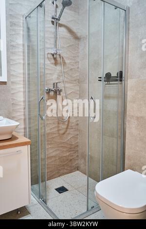 Questa immagine presenta un moderno box doccia in vetro in un bagno minimalista caratterizzato da piastrelle beige e eleganti finiture cromate, che mettono in risalto il contemporaneo Foto Stock