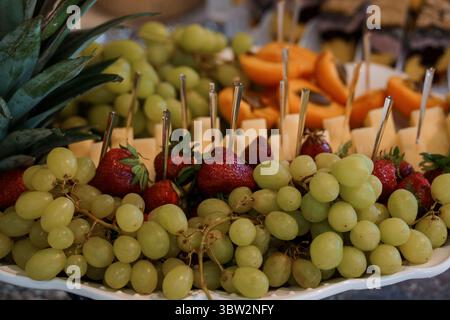Uno shot ravvicinato mostra un piatto di frutta ben organizzato con uva verde fresca, fragole mature, una fetta di ananas, cantaloupe e formaggi Foto Stock