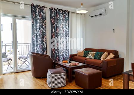 Un accogliente soggiorno con divano marrone, tende a motivi geometrici e accesso al balcone. il design degli interni e' moderno ed elegante, offrendo un ambiente accogliente Foto Stock