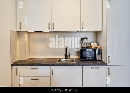 Una cucina moderna con armadi bianchi e un elegante ripiano nero, completo di elettrodomestici essenziali come un microonde e un bollitore. perfetto per Foto Stock