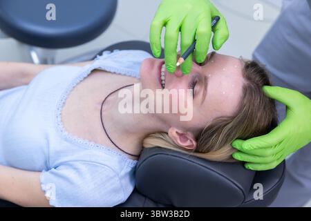 Primo piano di una procedura di bellezza: Una donna sorridente mentre riceve un trattamento cosmetico con un professionista che contrassegna la sua fronte in una clinica moderna Foto Stock