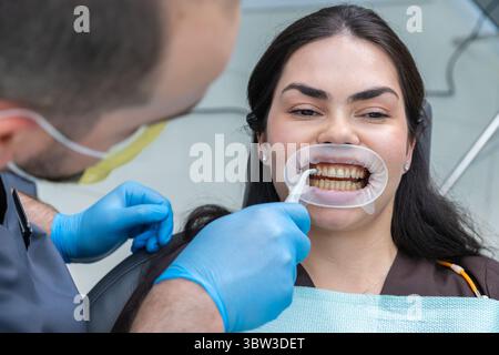 Paziente che riceve cure dentistiche mentre sorride, mostrando denti sani in un ufficio moderno da un dentista professionista con attrezzatura protettiva e strumenti per Foto Stock