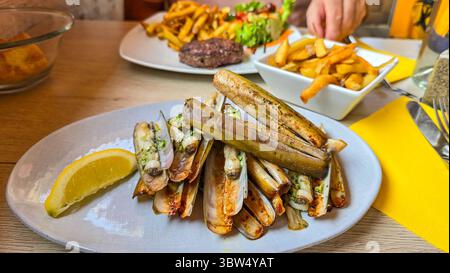 Appetitoso rasoio di spada con conchiglie fritte con aglio, prezzemolo e un pezzo di limone. Nelle vicinanze si trovano patate fritte Foto Stock