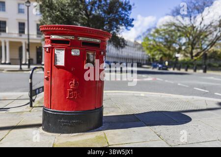Primo piano di una tradizionale cassetta postale rossa britannica su un marciapiede della città con cieli blu e alberi frondosi alle spalle. Londra, Regno Unito, 1° aprile 2024 Foto Stock