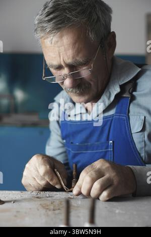 Un maestro anziano con gli occhiali al lavoro. Un vecchio bianco di aspetto caucasico in abiti da lavoro siede a un tavolo e lavora con le mani Foto Stock