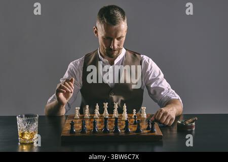 Barbuto bella uomo con un elegante baffi, vestito con un classico giubbotto marrone e camicia bianca è la localizzazione a un tavolo e giocare a scacchi byhimself, lo Foto Stock