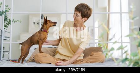 Proprietaria che gioca con un cane gioioso a casa, una donna matura felice che si diverte ad allenarsi con il suo simpatico pinscher in miniatura. Giocare con il concetto di cane a Hom Foto Stock