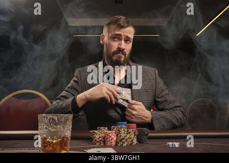 Bel maschio barbuto in un classico vestito grigio sta giocando a poker al casinò in fumo seduto al tavolo con patatine e carte sopra. Nasconde un po' di c Foto Stock