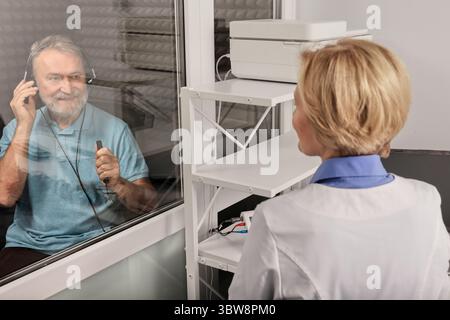 Esame audiologico. Audiologo che fa check-up uditivo a un uomo anziano in cabina audiometrica insonorizzata. Trattamento perdita dell'udito, vista laterale Foto Stock