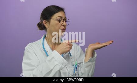 Medico donna che indossa un cappotto bianco e un dito dello stetoscopio che punta sul palmo aperto in studio viola; sicurezza. Foto Stock
