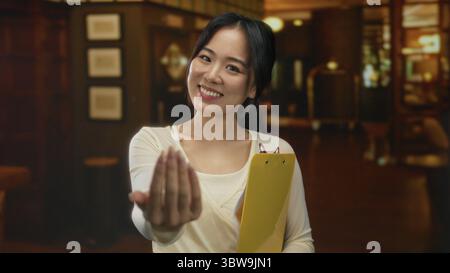 Giovane donna cinese in un hotel sorridente con fiducia tenendo un appunti giallo all'interno, catturando un gesto caldo e accogliente in un ambiente professionale. Foto Stock
