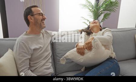 Coppia ispanica di mezza età che ride insieme su un comodo divano di casa in un luminoso soggiorno, catturando l'amore e i momenti di gioia in casa. Foto Stock