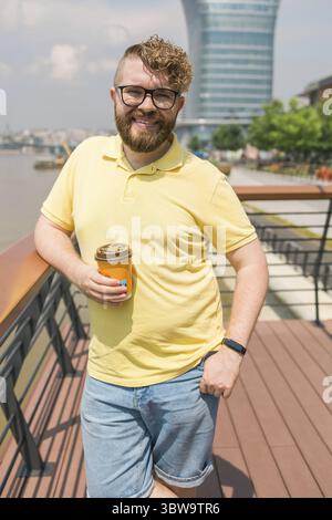 Un giovane uomo giovane e attraente con i capelli ricci che indossa gli occhiali, cammina per strada con una tazza di caffè in una giornata di sole. Un hipster barbuto che indossa Foto Stock